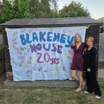 two women smiling in front a hand painted banner saying 'blakeney house 20 years'